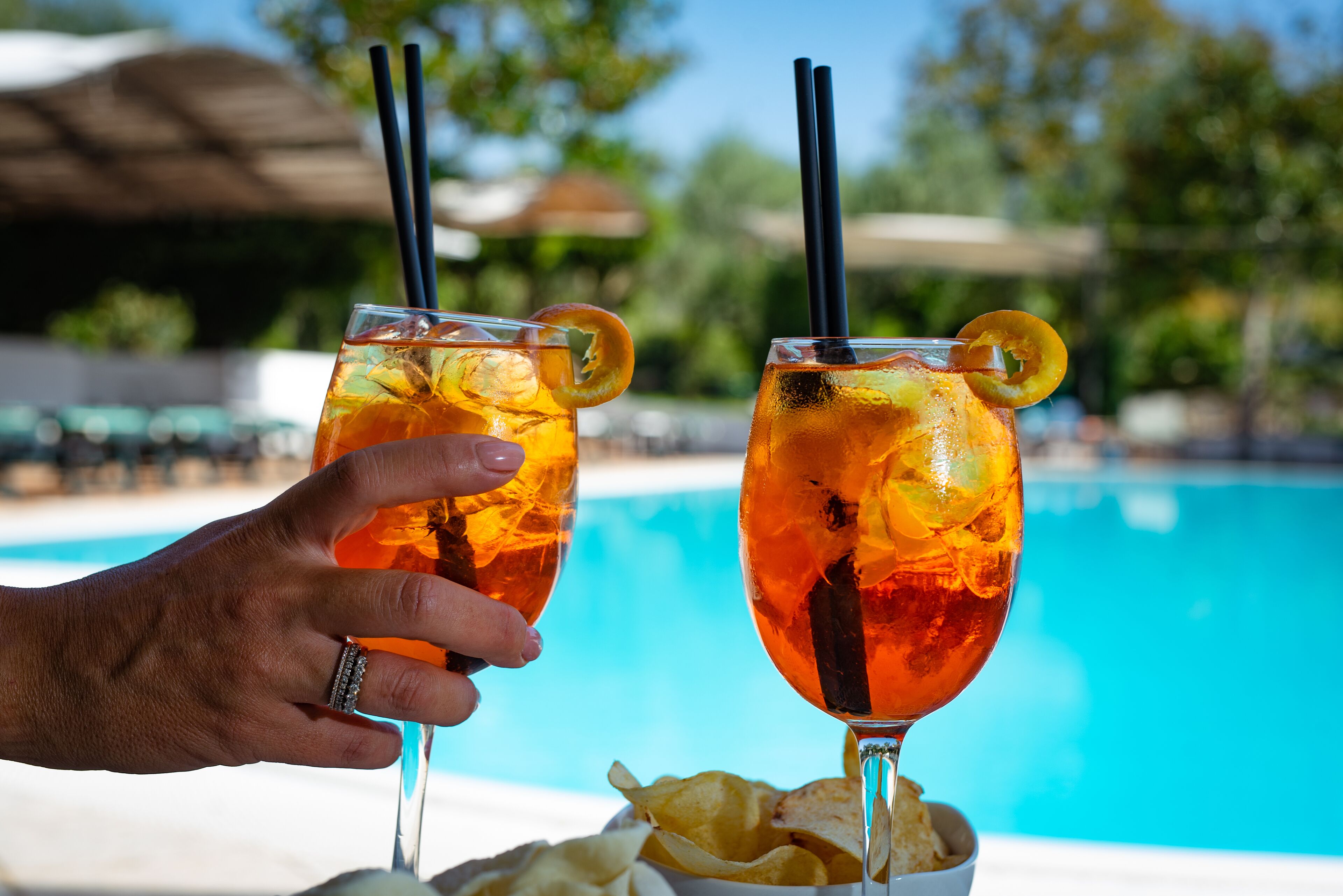 Two cocktails with lemon slices and straws, a pool in the background, and a hand holding one of the drinks.