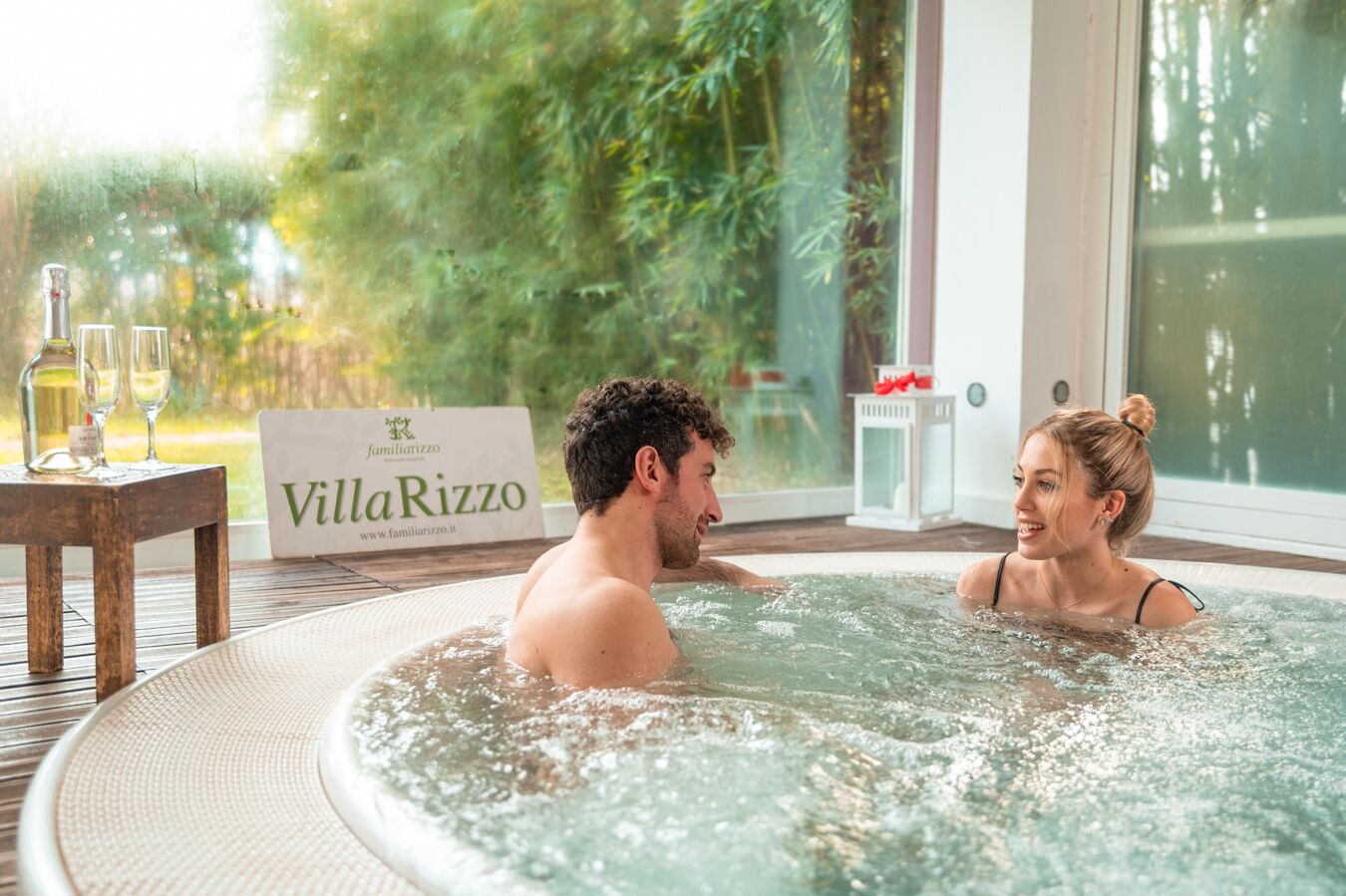 Ett par sitter i ett bubbelbad, och en skylt med texten ”VillaRizzo” syns i det väl upplysta rummet. Utanför kan man se grönskan.