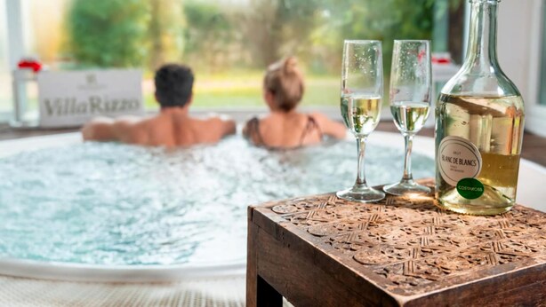 A hot tub with two people, a bottle of white wine, and two glasses on a wooden table. - Hotel Villa Rizzo Resort & Spa (San Cipriano Picentino)