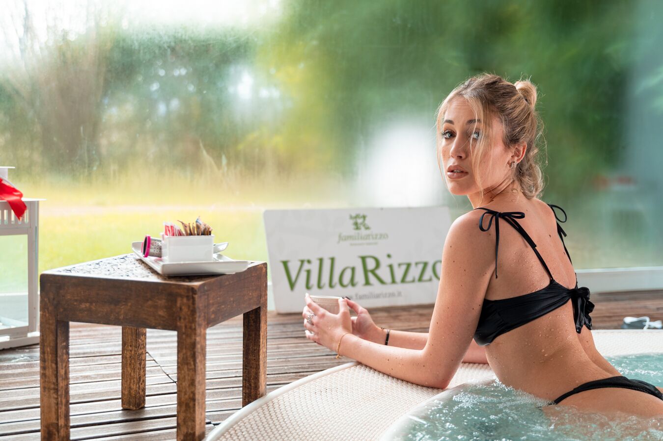 Una persona con traje de baño negro sentada en un jacuzzi con una mesa de madera y un cartel que dice "VillaRizzo" al fondo.