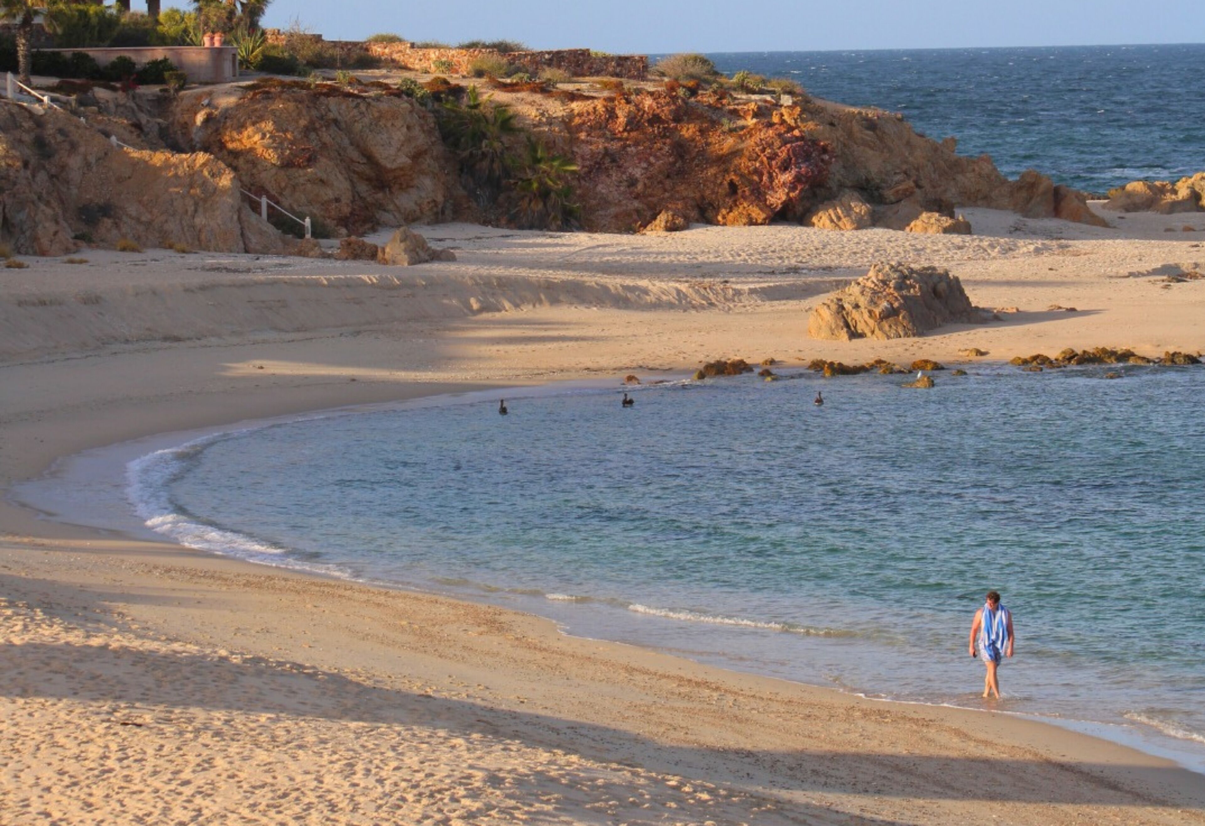 A beach with a rocky cliff, clear water, and a person walking.
