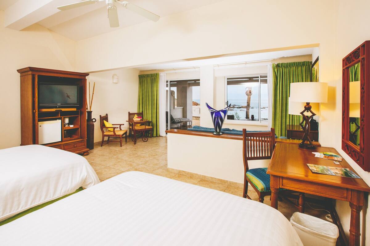 A hotel room with two beds, a television, a dining table, a lamp, and a view of the sea.