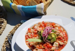 Un plato blanco con un plato de mariscos, posiblemente ceviche, adornado con aguacate y cebollas rojas, acompañado de una cesta de pan y una servilleta azul.