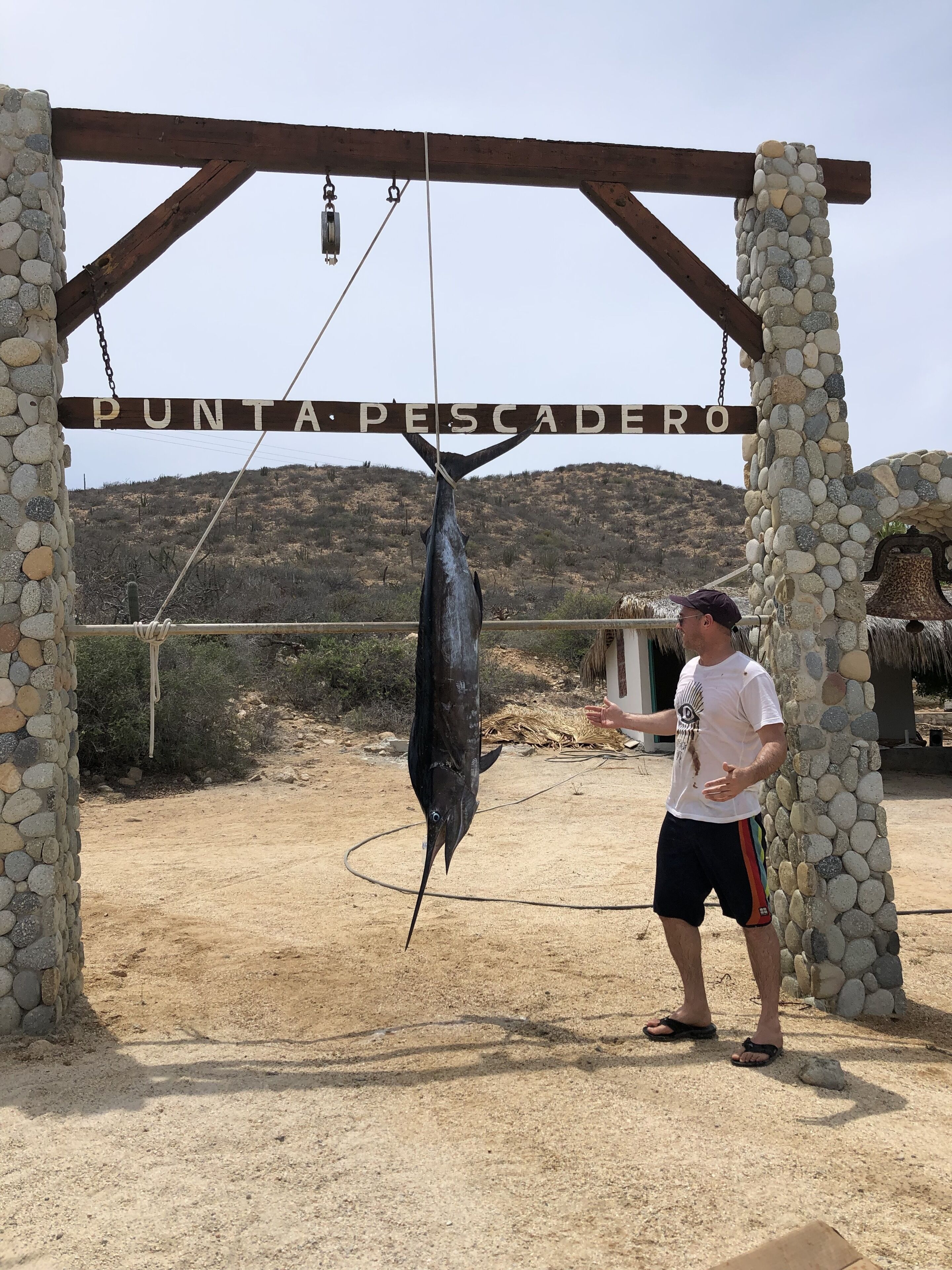 A man standing under a wooden archway with the sign "Punta Pescadero".