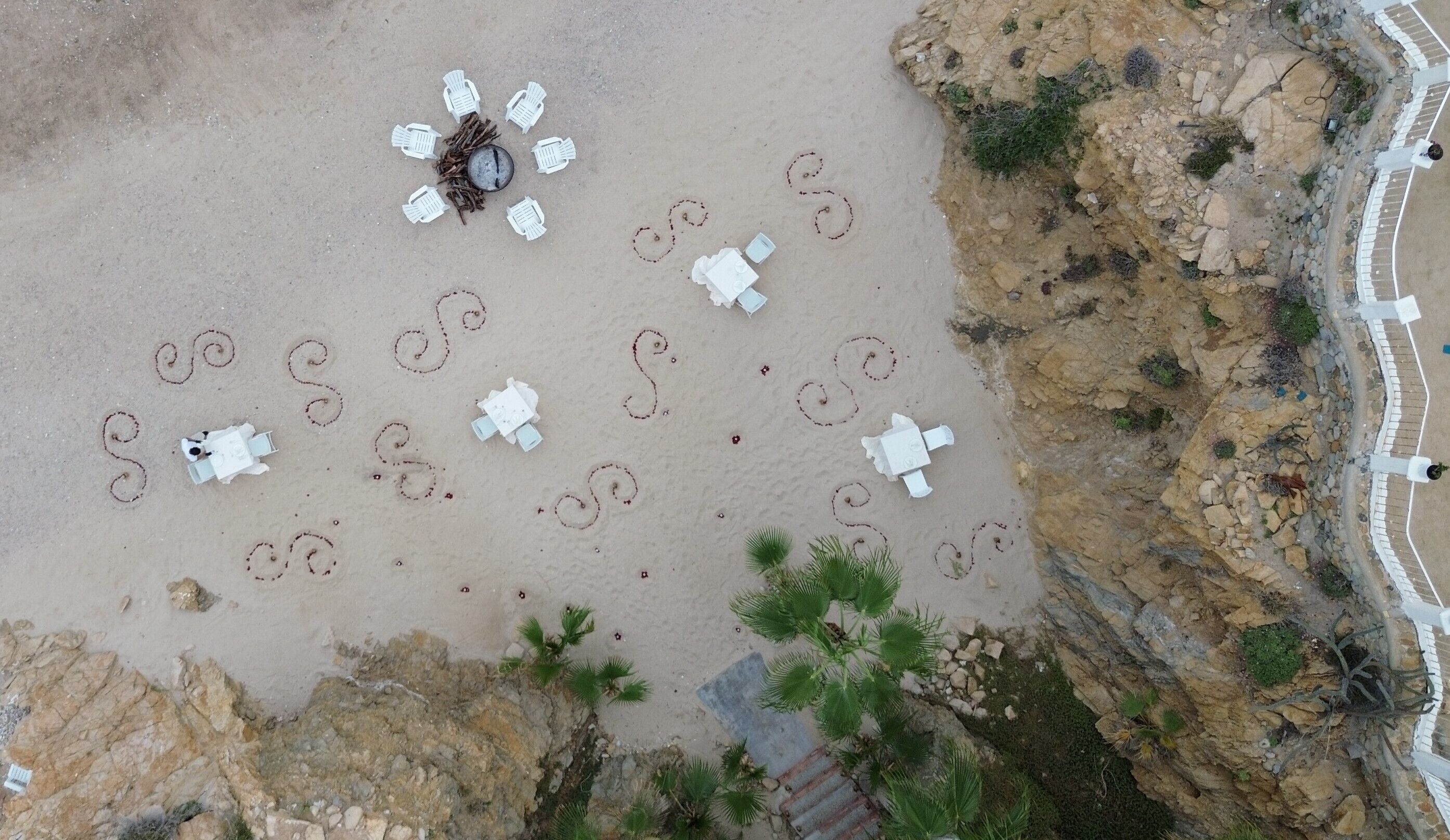 Vista aérea de una playa con sillas de descanso blancas dispuestas en círculo alrededor de una zona central con mesa y sillas.
