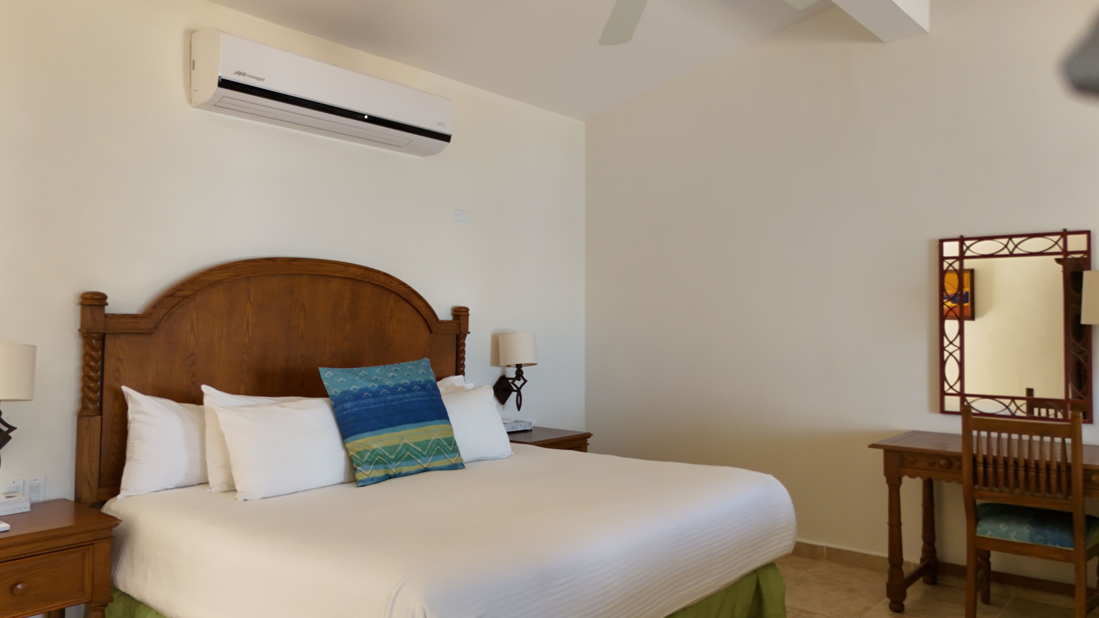 Une chambre avec un lit en bois, une literie blanche, une table de chevet avec une lampe, une commode, un miroir et un appareil de climatisation fixé au mur.