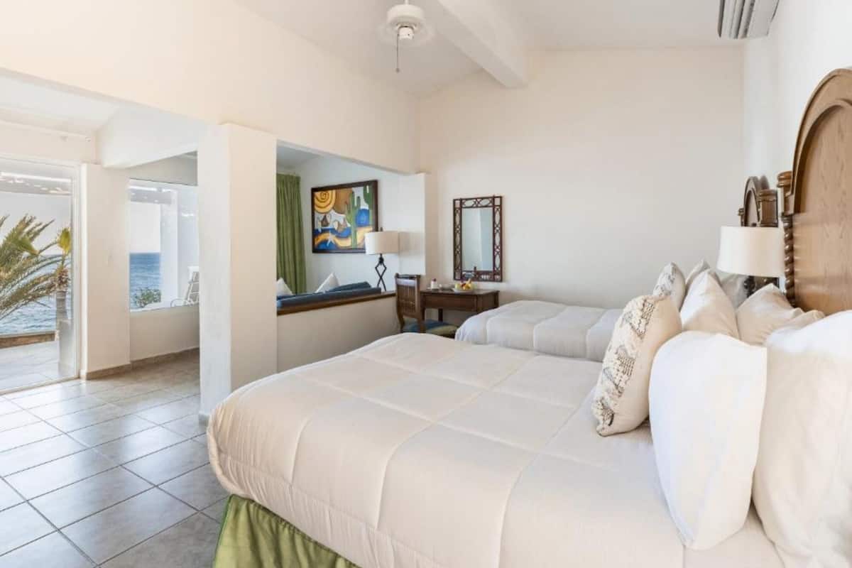 A hotel room with a large bed, a desk with a chair, a mirror, and a view of the sea.