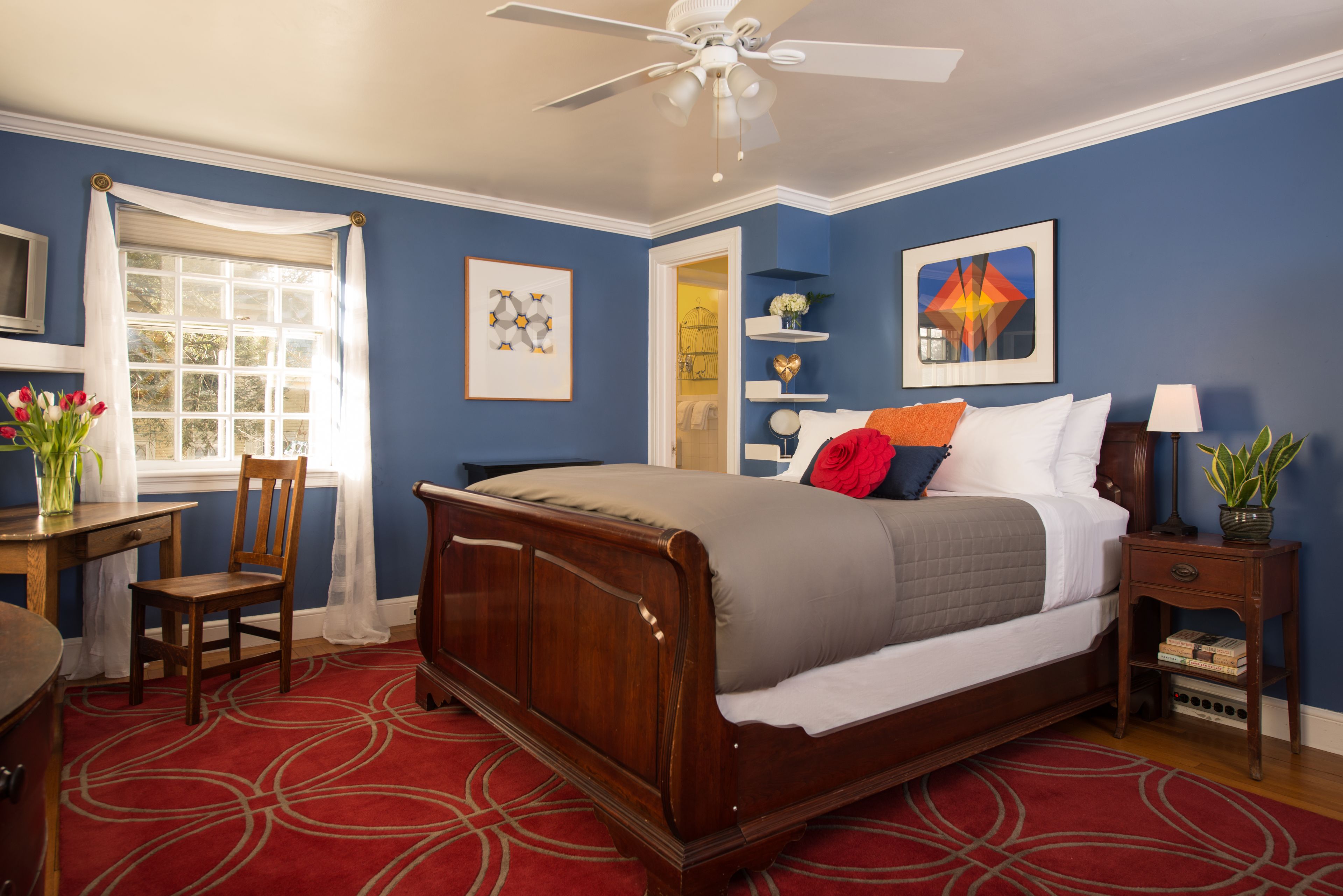 Quarto com cama de madeira, tapete vermelho, ventilador de teto e janela com cortinas brancas.