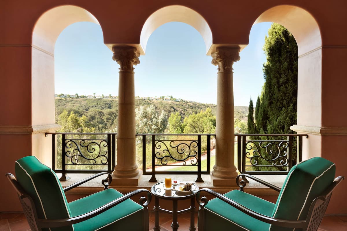 A balcony with two green upholstered chairs and a small table with a glass of orange juice, overlooking a hilly landscape with trees and a clear sky.