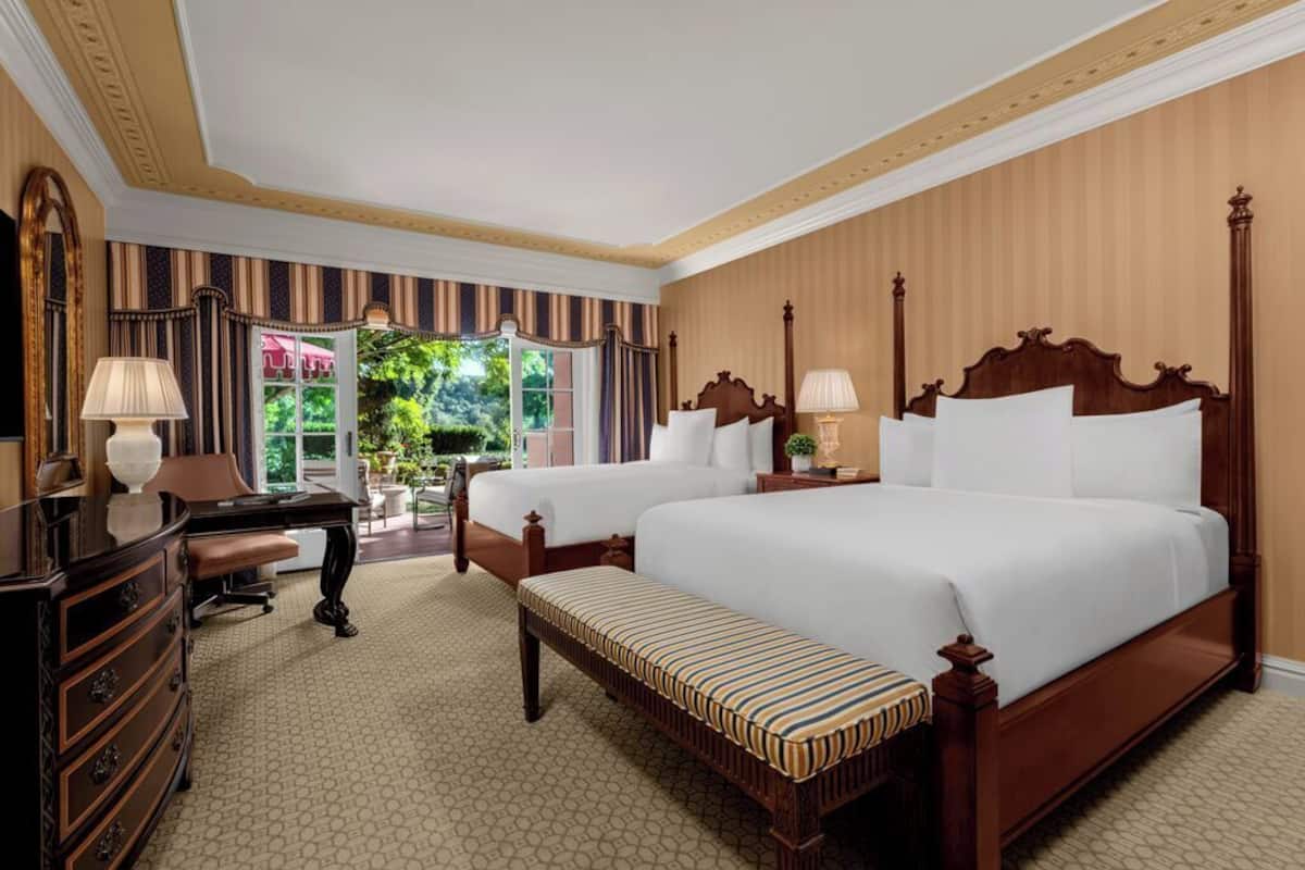 A hotel room with two beds, a desk, a dresser, and a bench.