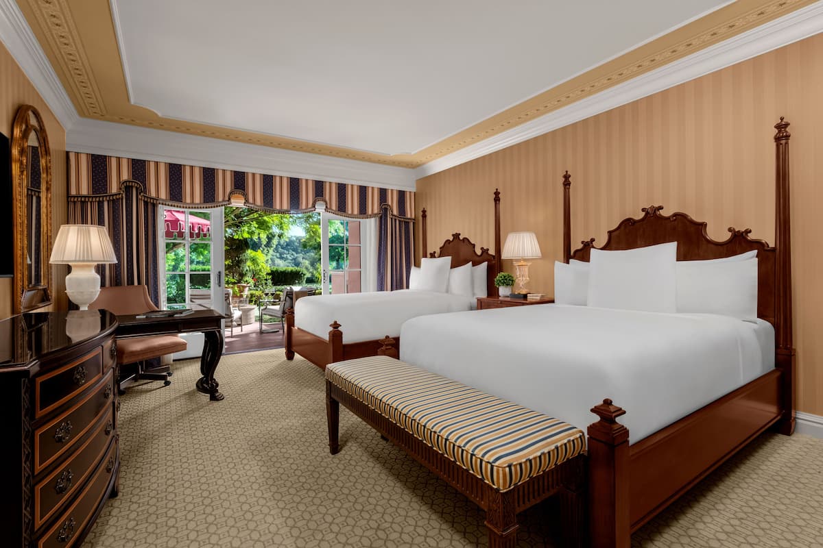 A hotel room with two beds, a desk, a chair, a dresser, and a window with a view of greenery.
