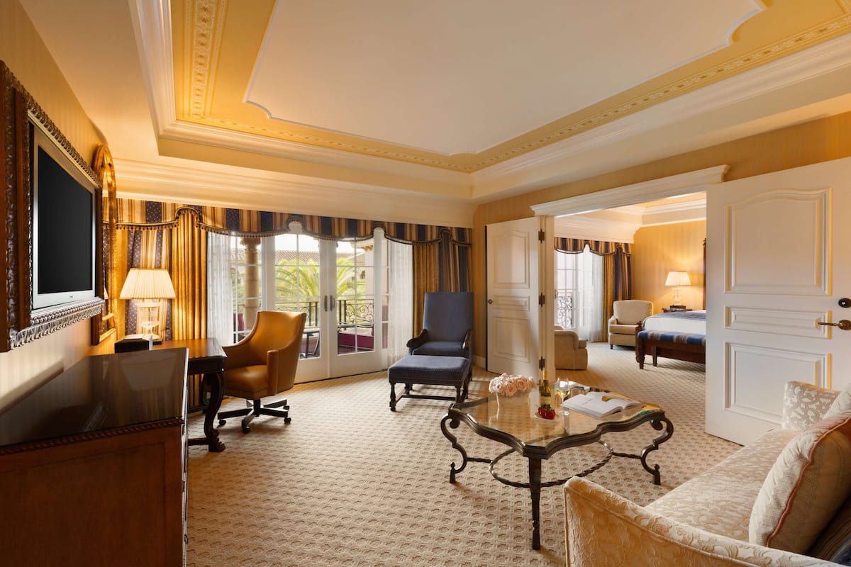 A hotel room with a large bed, a sitting area with a sofa and armchairs, a dining table, and a TV.