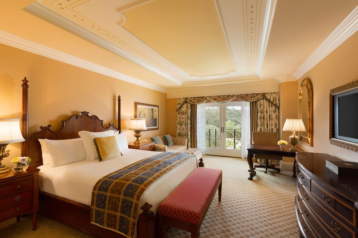 A hotel room with a large bed, a bench, a desk, a television, and a balcony with curtains.