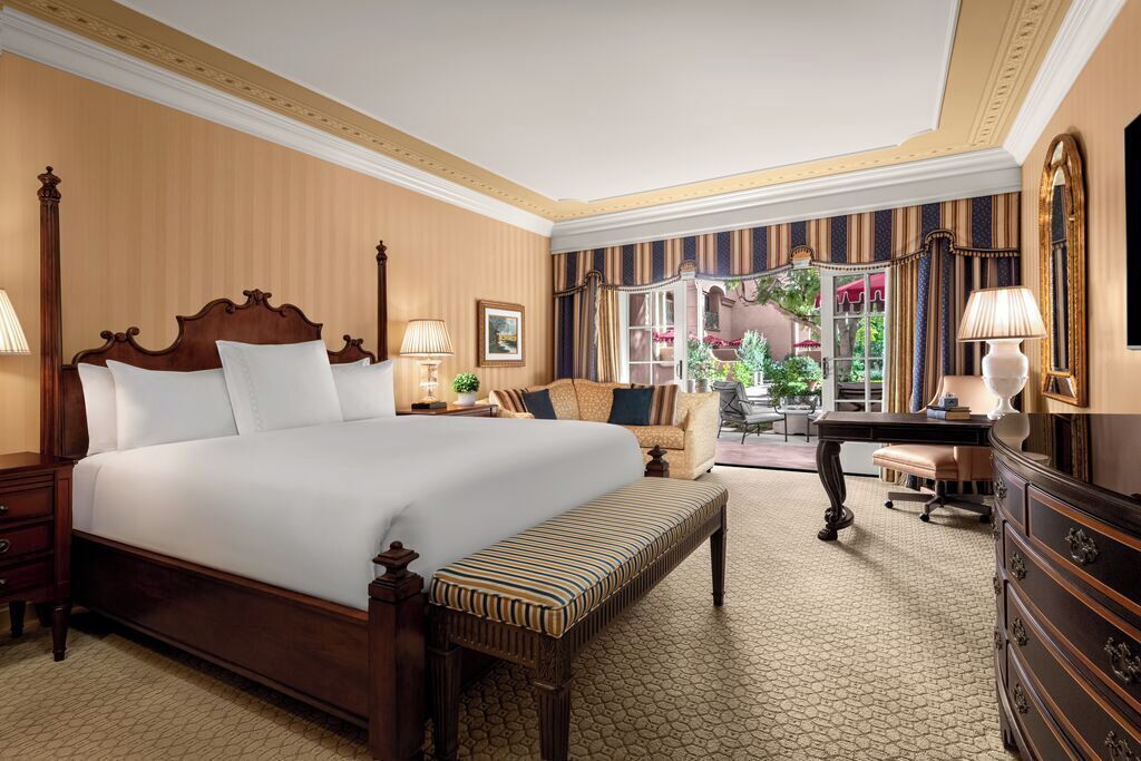 A hotel room with a large bed, a desk, a chair, and a bench. There is a window with a view of the outside.