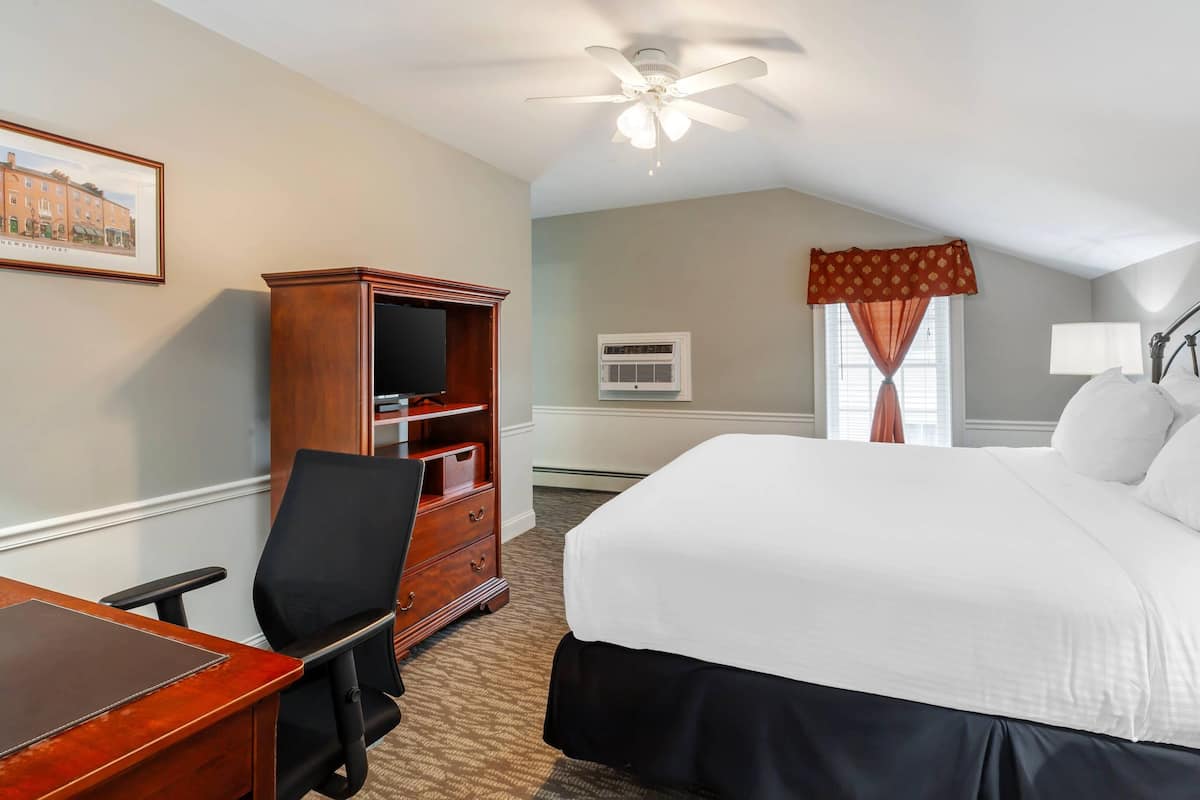 A hotel room with a large bed, a desk with a chair, a wooden wardrobe, a ceiling fan, an air conditioning unit, and a window with curtains.