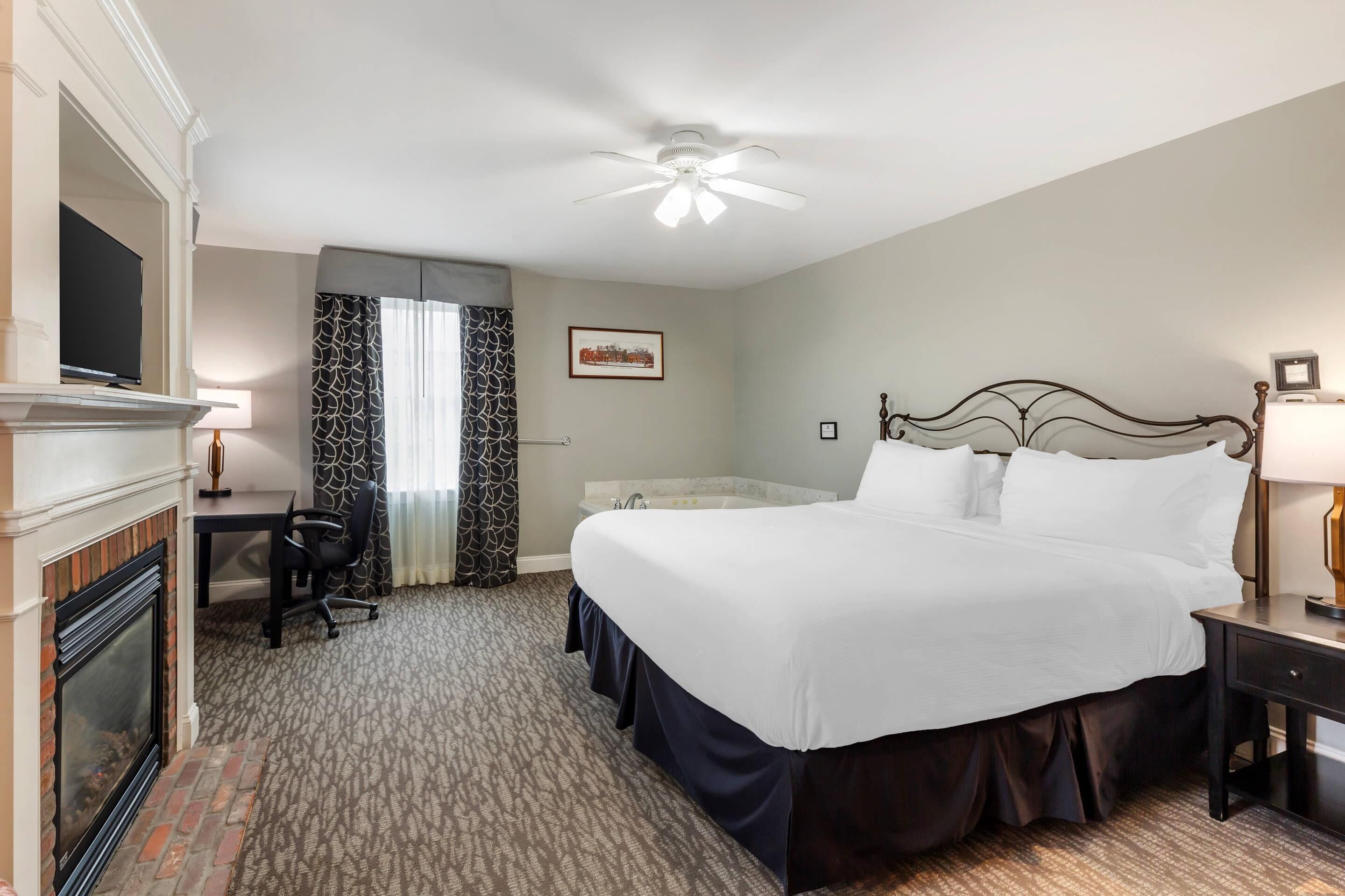 A hotel room with a large bed, a desk, a fireplace, and a ceiling fan.