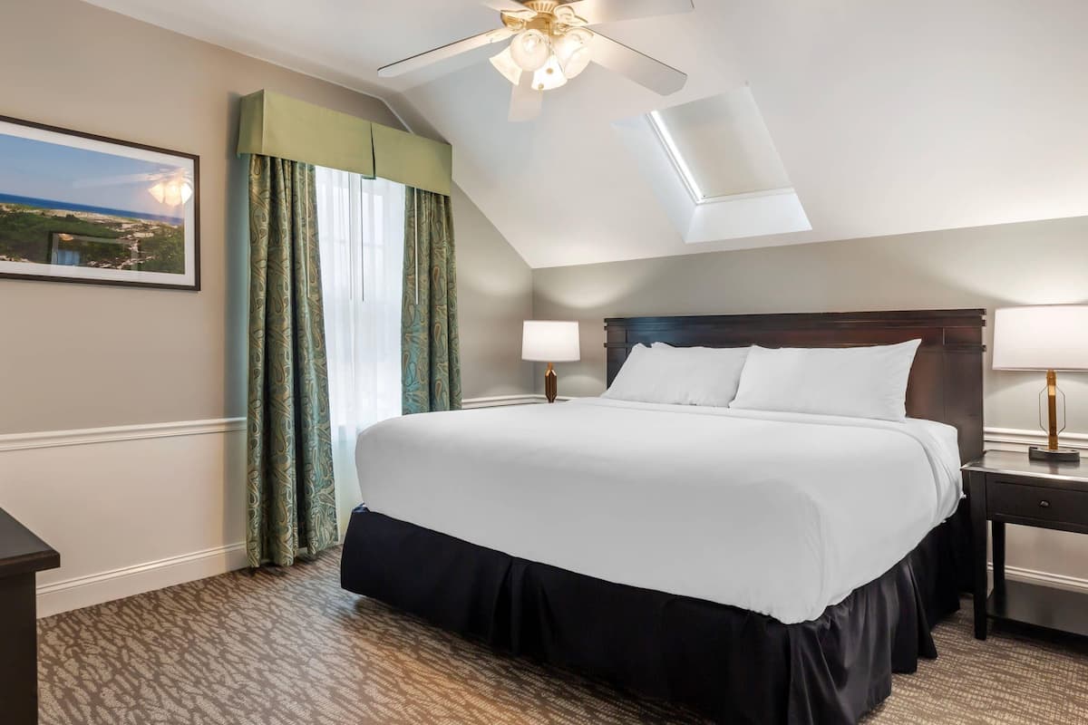 A hotel room with a large bed, a ceiling fan, a nightstand with a lamp, and a window with curtains.