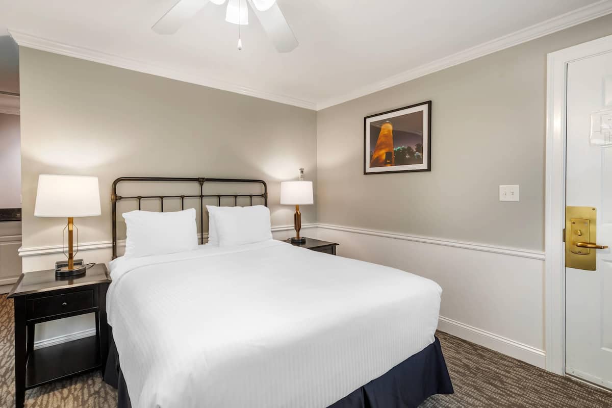 A neatly made bed with white linens, two bedside tables with lamps, a framed picture on the wall, and a door with a gold handle.
