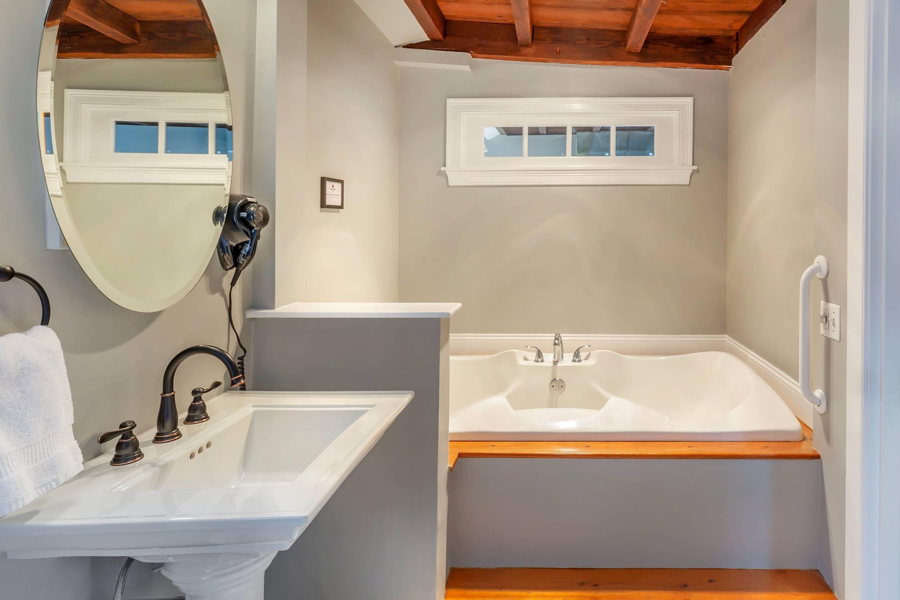 Une salle de bain avec un lavabo blanc, un grand miroir, une baignoire et une fenêtre au-dessus du lavabo.