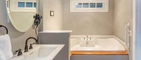 Une salle de bain avec un lavabo blanc, un grand miroir, une baignoire et une fenêtre au-dessus du lavabo.