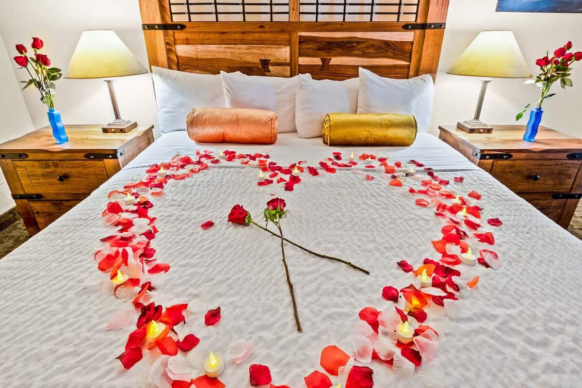 A neatly made bed with rose petals arranged in a heart shape and a single rose on the bed.