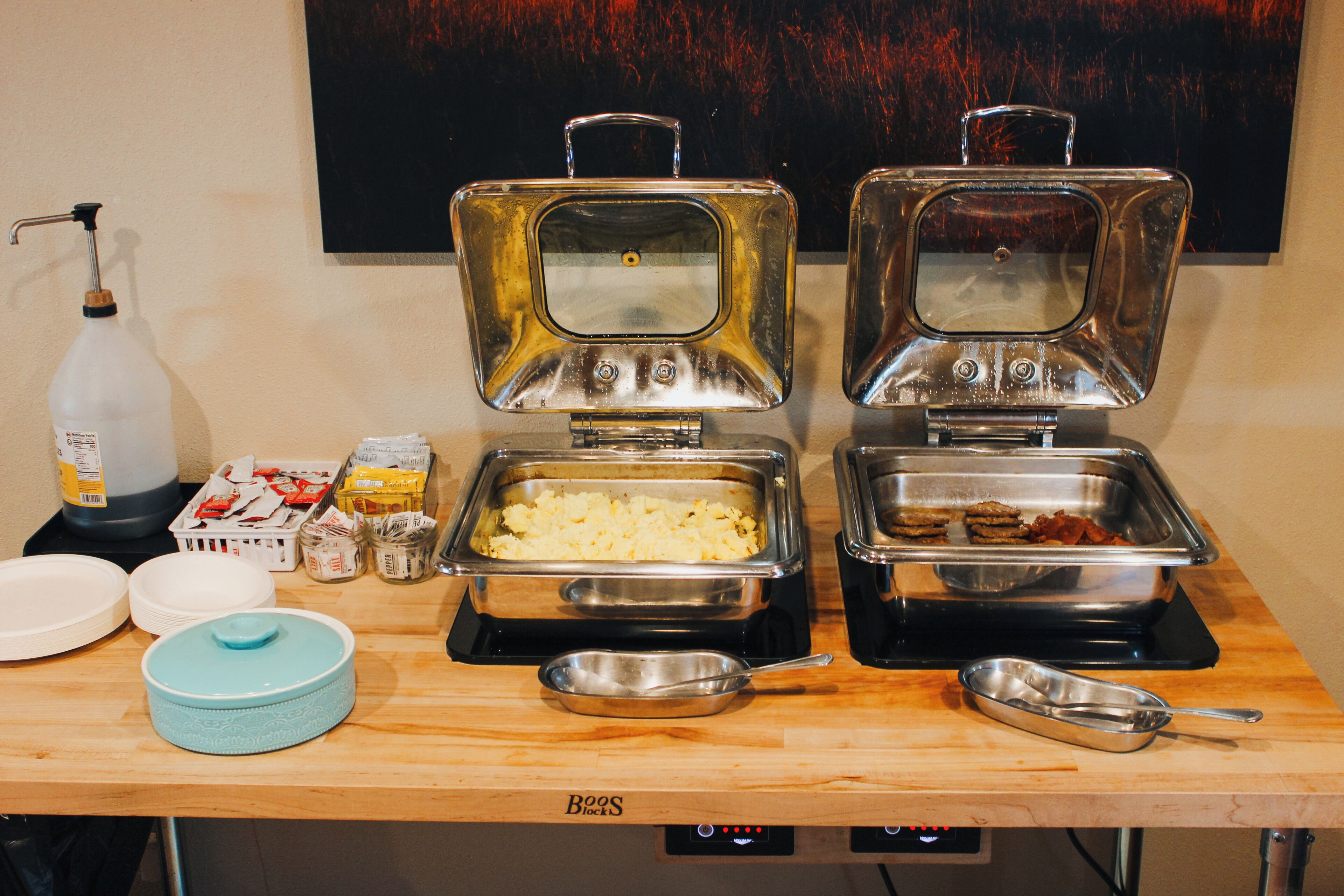 A buffet setup with two open food dispensers, a water dispenser, and various food containers on a wooden table.