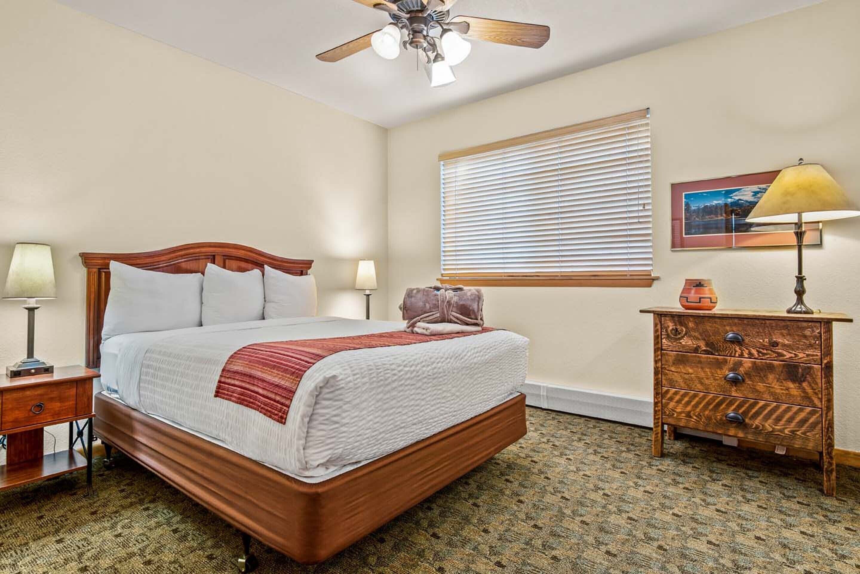 A neatly arranged bedroom with a wooden bed, a bedside table with lamps, a dresser, a ceiling fan, and a window with blinds.