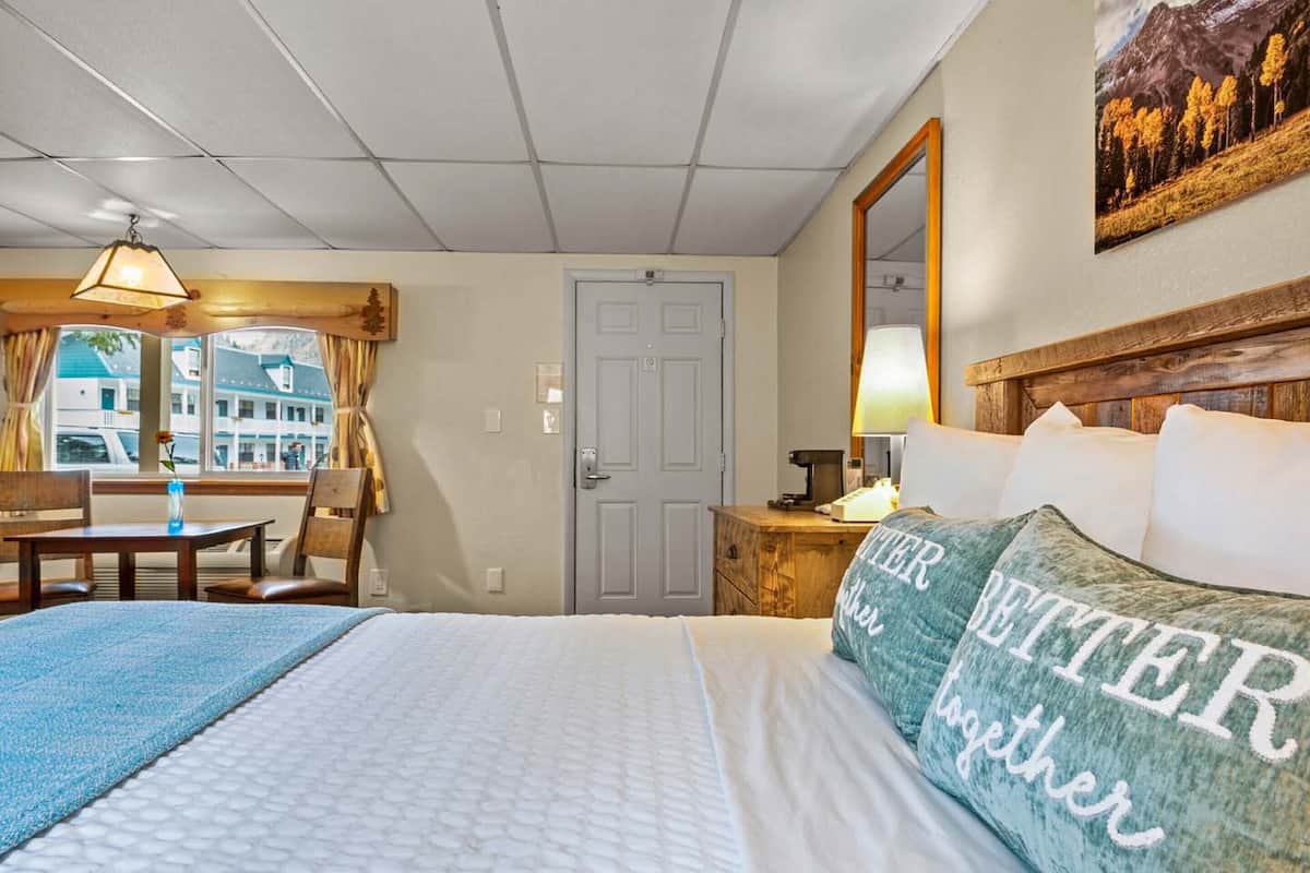 A hotel room with a bed, a desk, a chair, a window with curtains, a door, and a wall-mounted mirror.