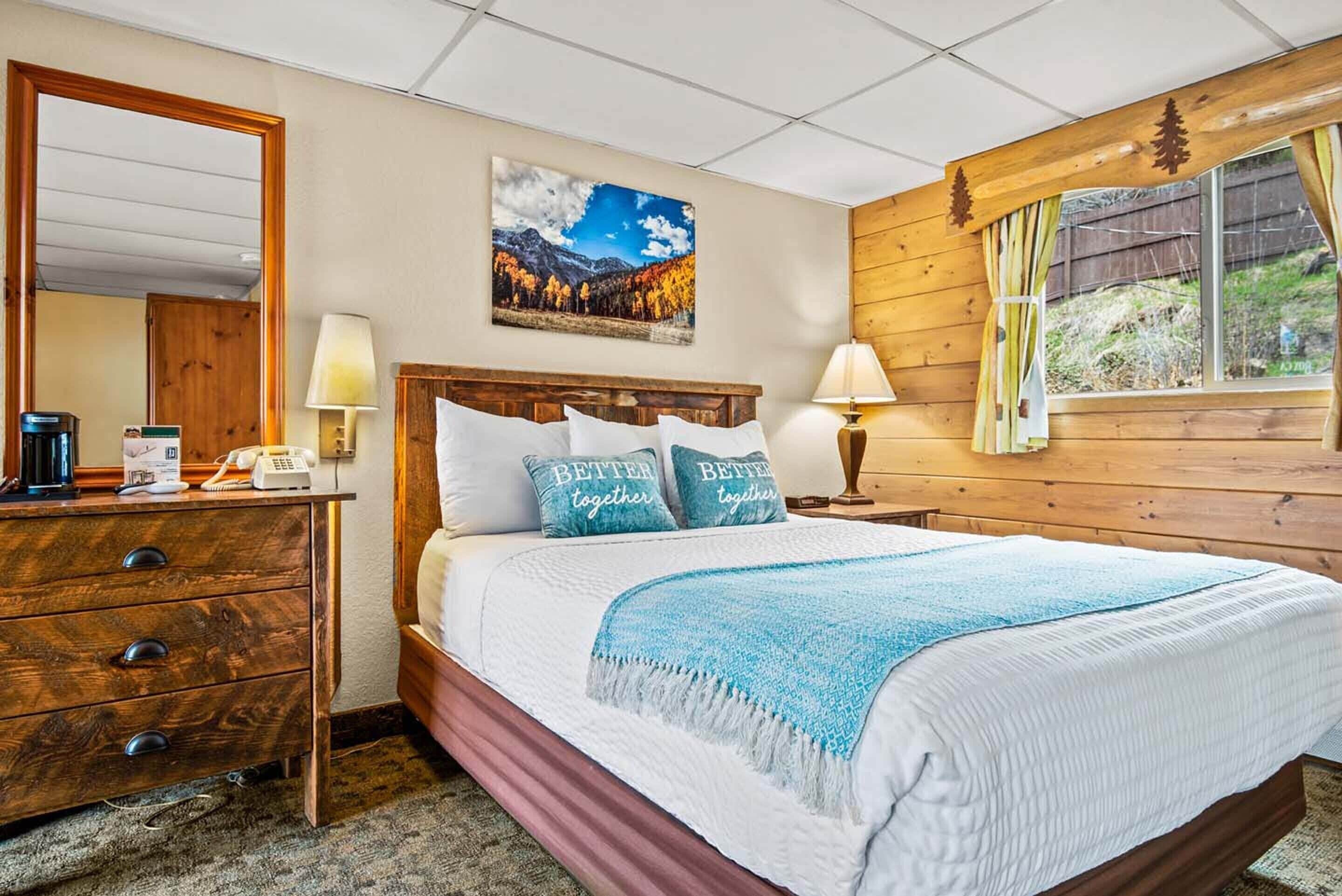 A bedroom with a wooden headboard, a bed with white bedding and blue pillows, a nightstand with a lamp, a dresser with drawers, a painting on the wall, and a window with curtains.