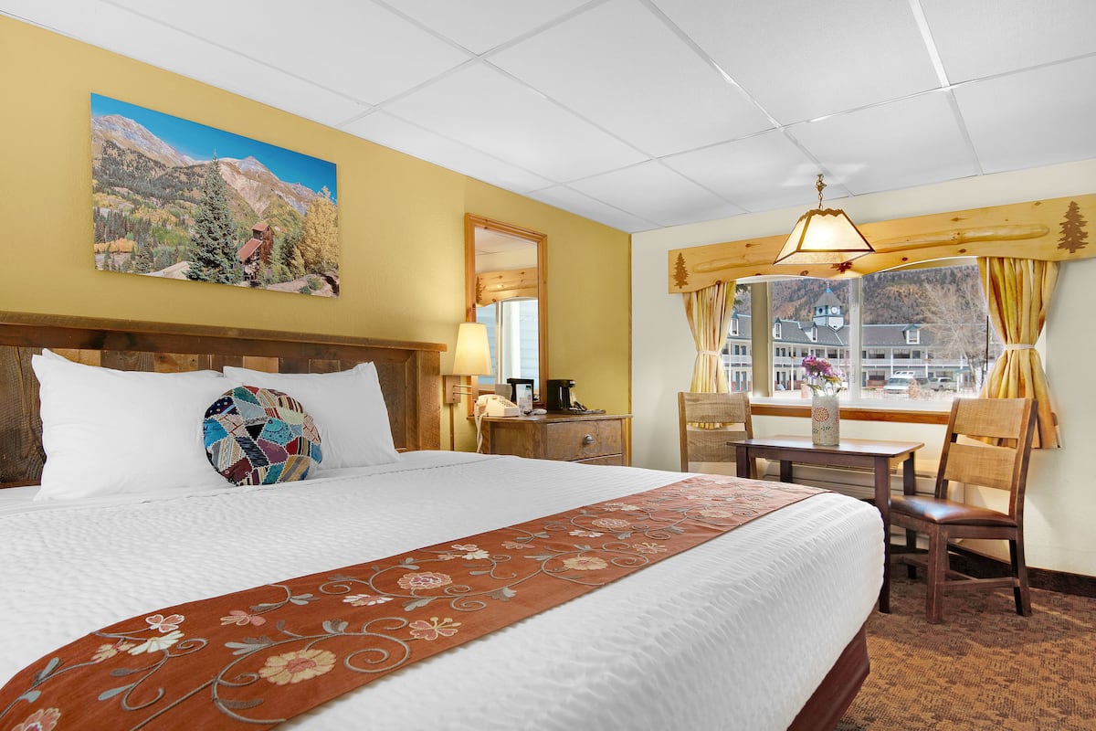 A hotel room with a large bed, a wooden headboard, a painting of a mountain landscape, a desk with a lamp, and a view of a town through the window.