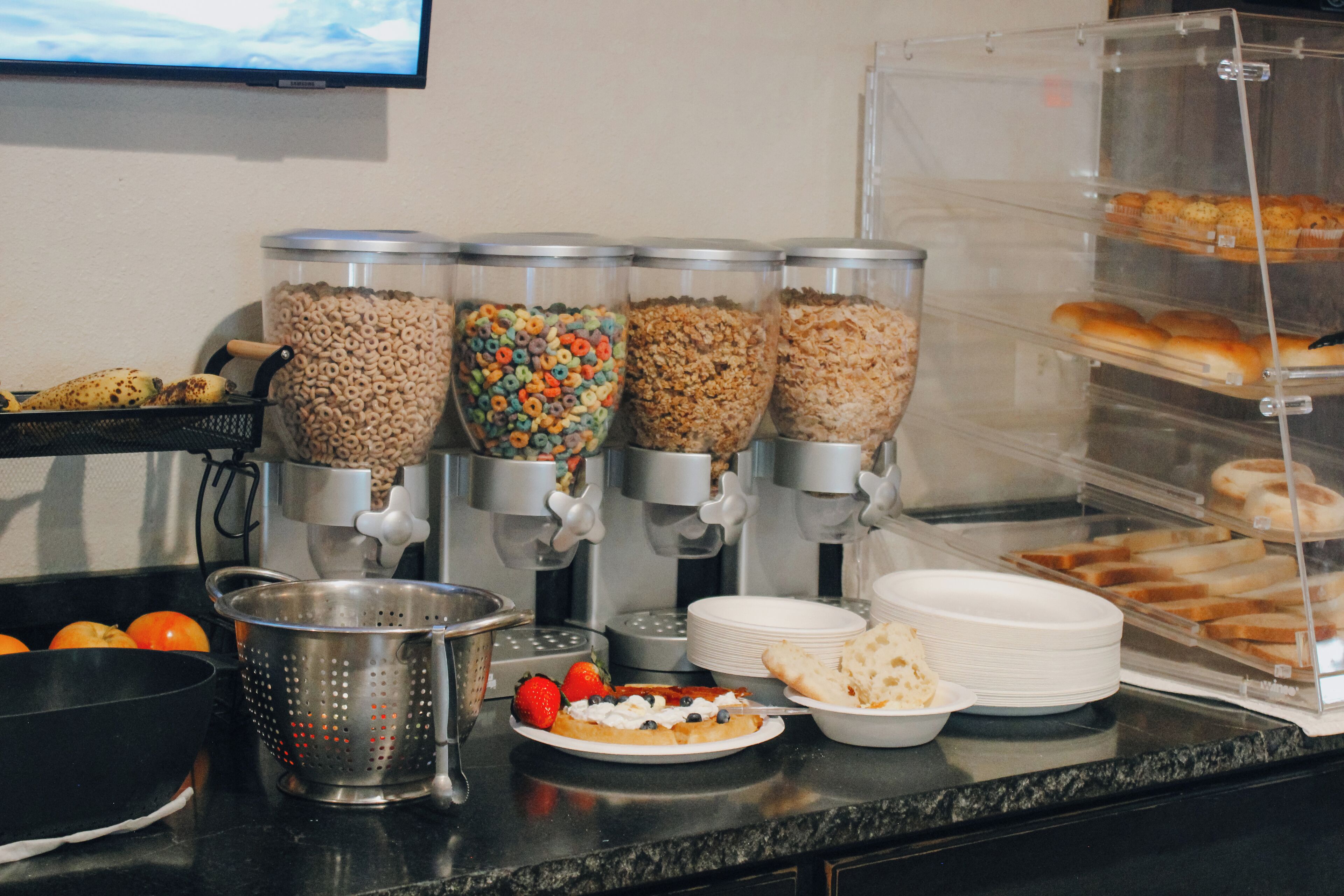 A buffet setup with cereal dispensers, a colander, plates, and a bread display.