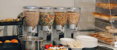 A buffet setup with cereal dispensers, a colander, plates, and a bread display.