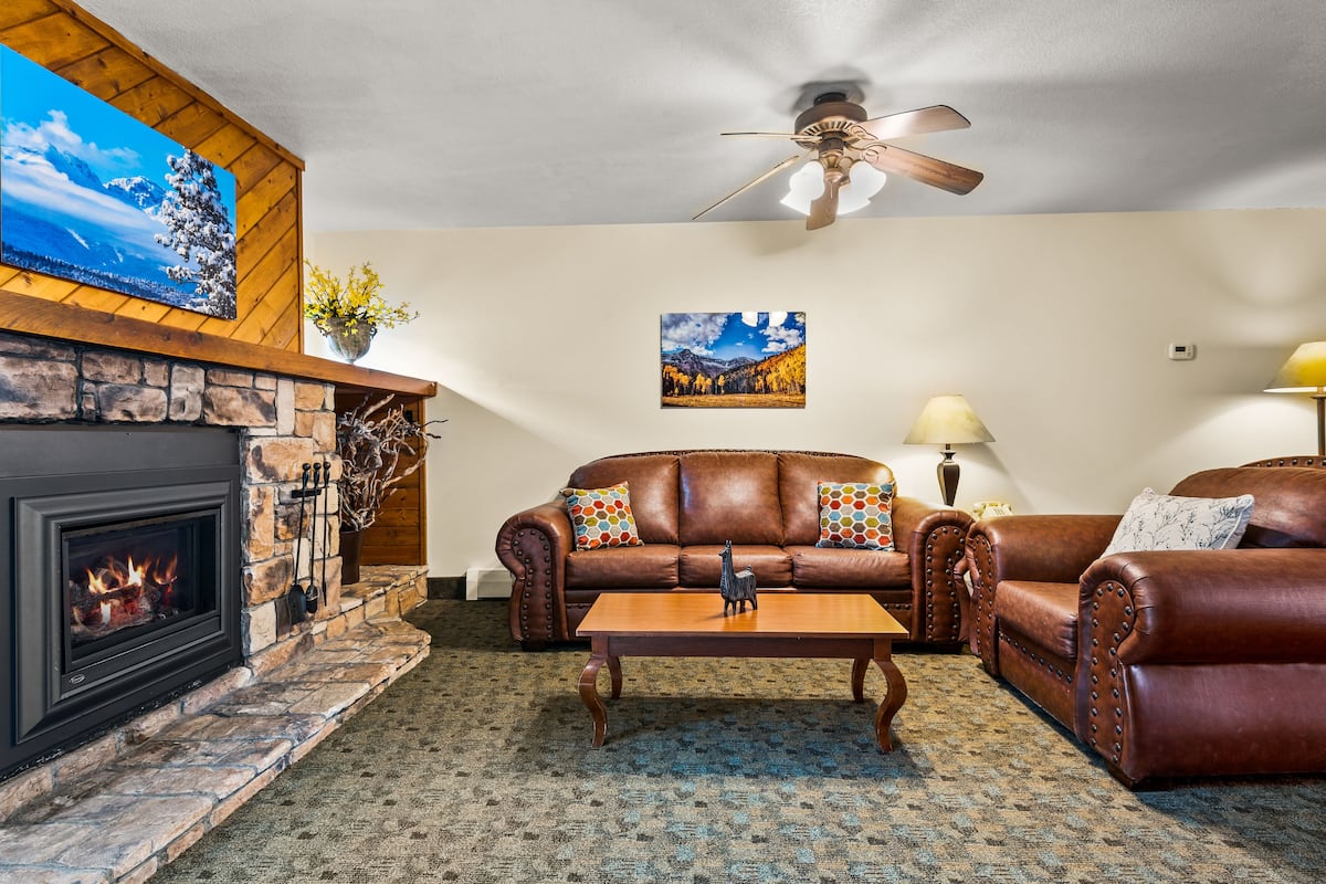 A cozy living room with a fireplace, leather sofas, a coffee table, and a ceiling fan.