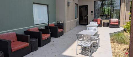 An outdoor seating area with wicker furniture and metal chairs, a tiled floor, and a building entrance with glass doors.