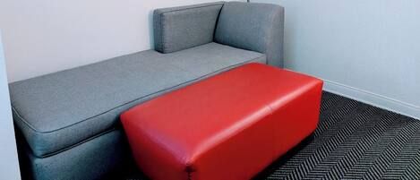 Una habitación con un sofá gris, un reposapiés rojo, un cuadro abstracto enmarcado y una lámpara.