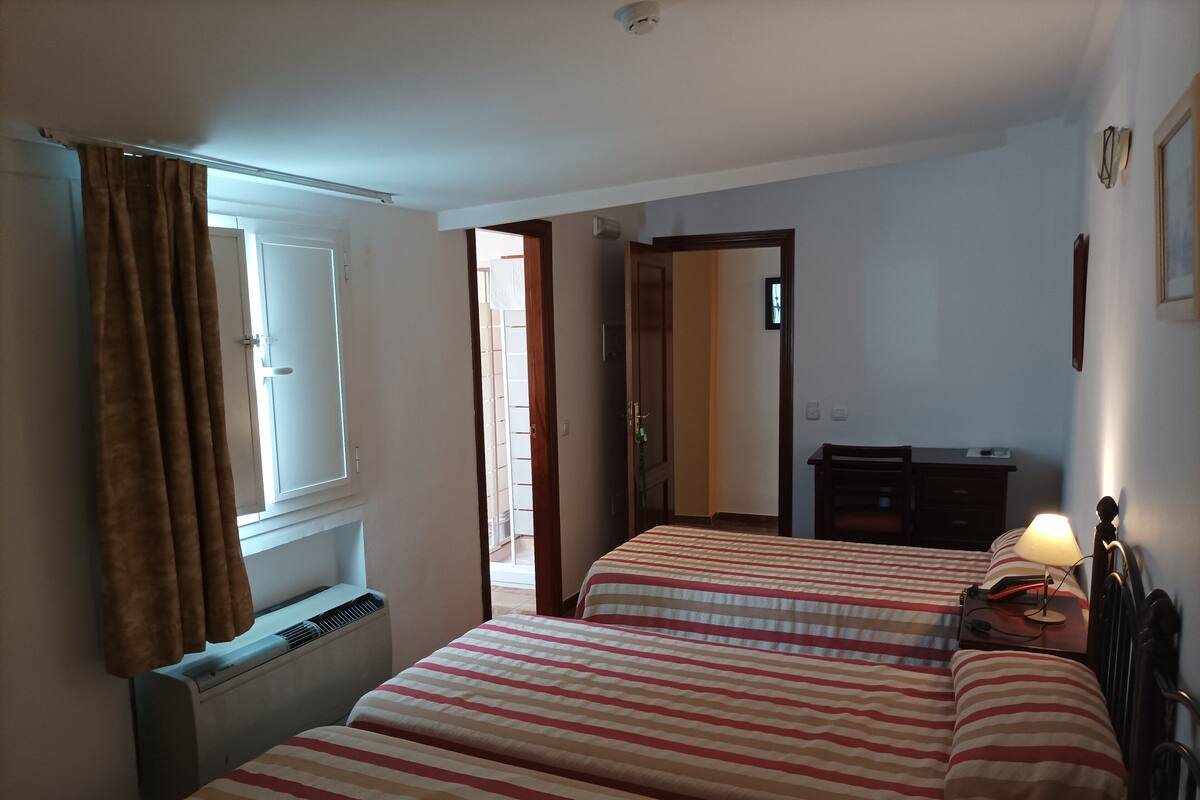 Une chambre avec deux lits, une fenêtre avec des rideaux, un radiateur, un bureau avec une lampe et une porte donnant sur une autre pièce.
