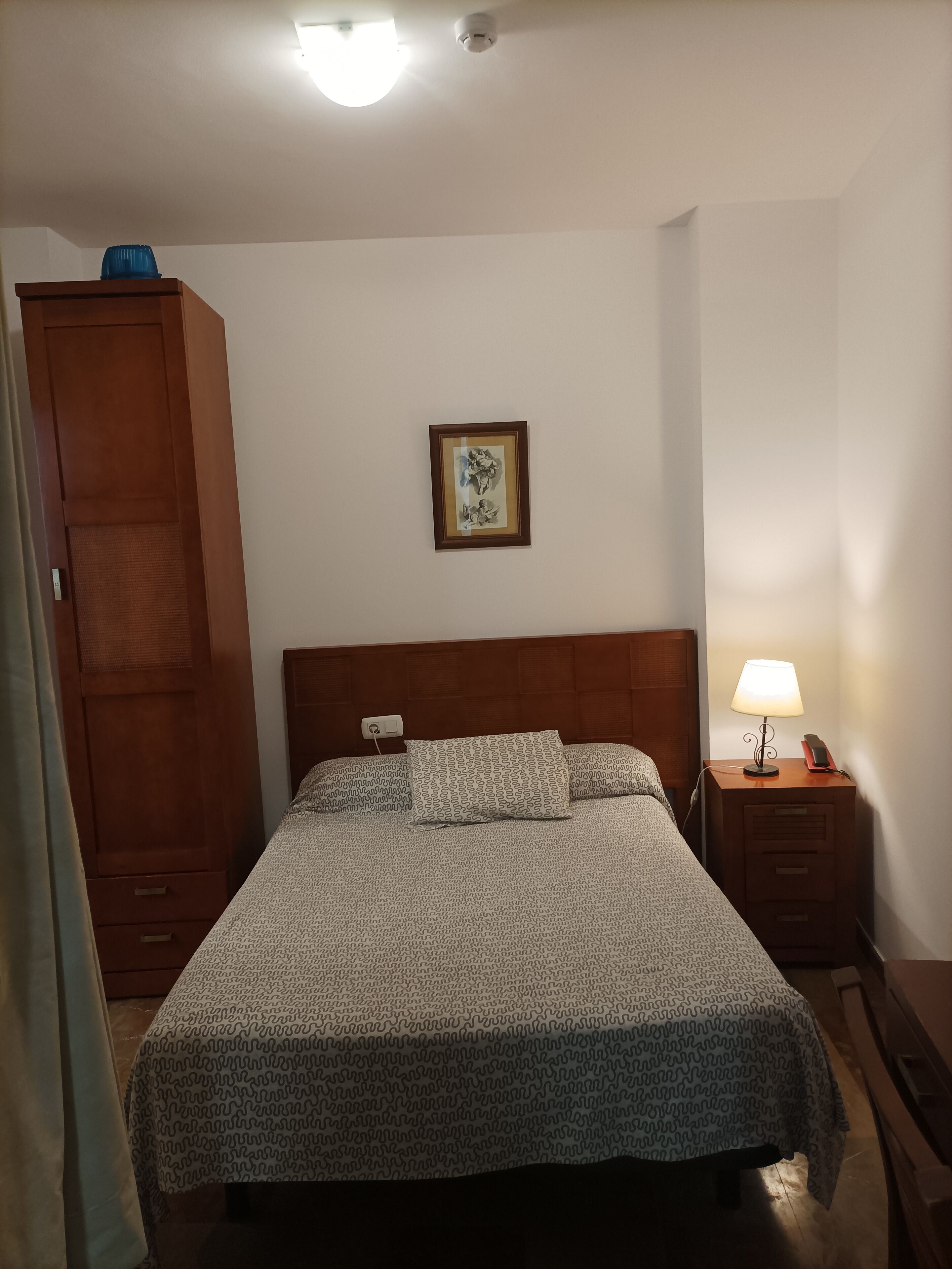 Een slaapkamer met een bed, nachtkastje met lamp, kledingkast en een lijst aan de muur.