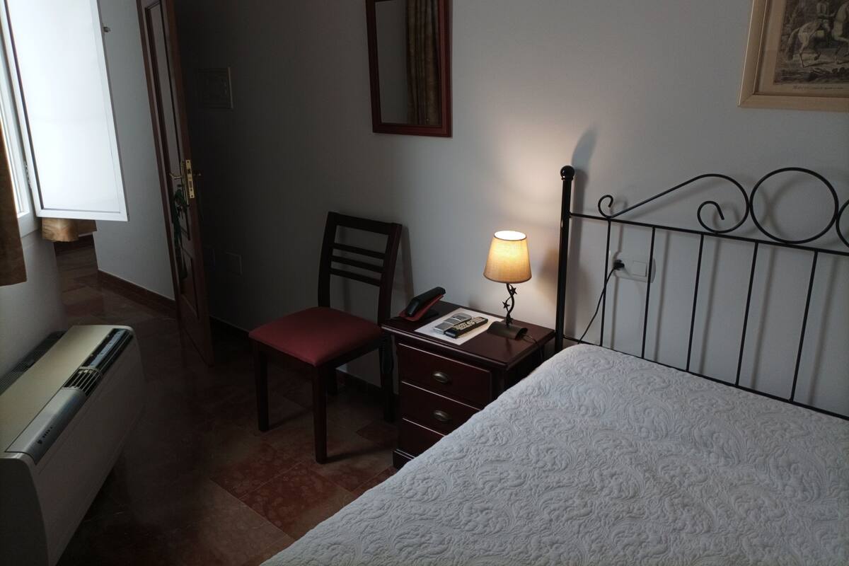 Une chambre d’hôtel avec un lit, une chaise, un bureau avec une lampe, un téléphone et un radiateur.