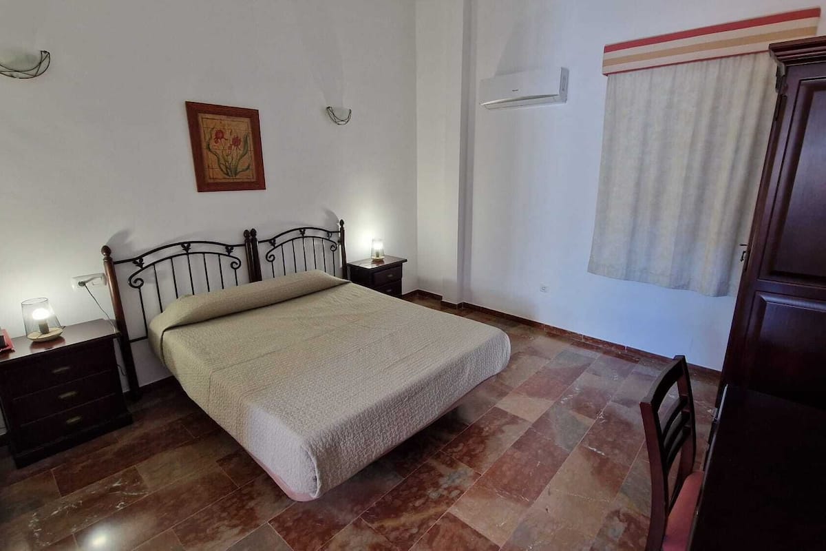 Une chambre à coucher comprenant un lit, des tables de chevet, une commode, une armoire en bois, une fenêtre avec des rideaux et un tableau accroché au mur.