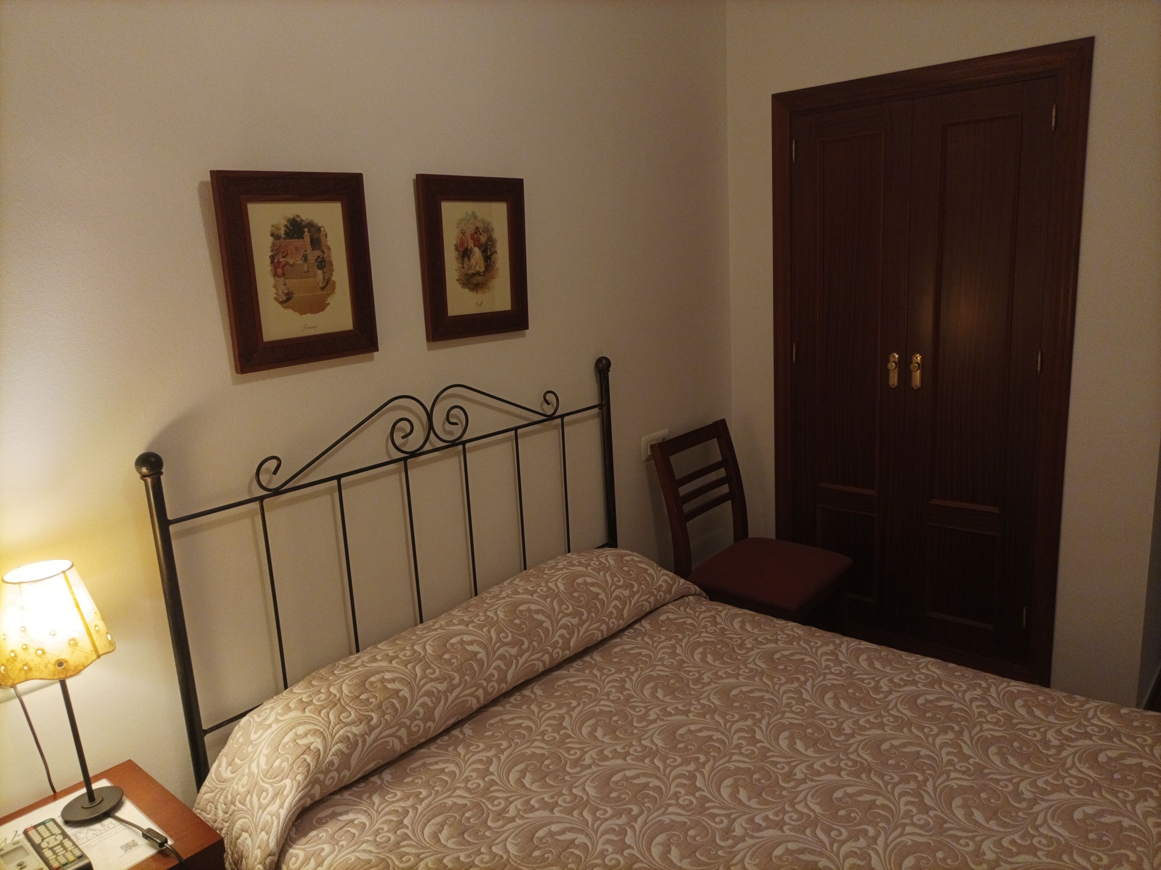A neatly made bed with a patterned bedspread, a bedside table with a lamp, and two framed pictures on the wall.
