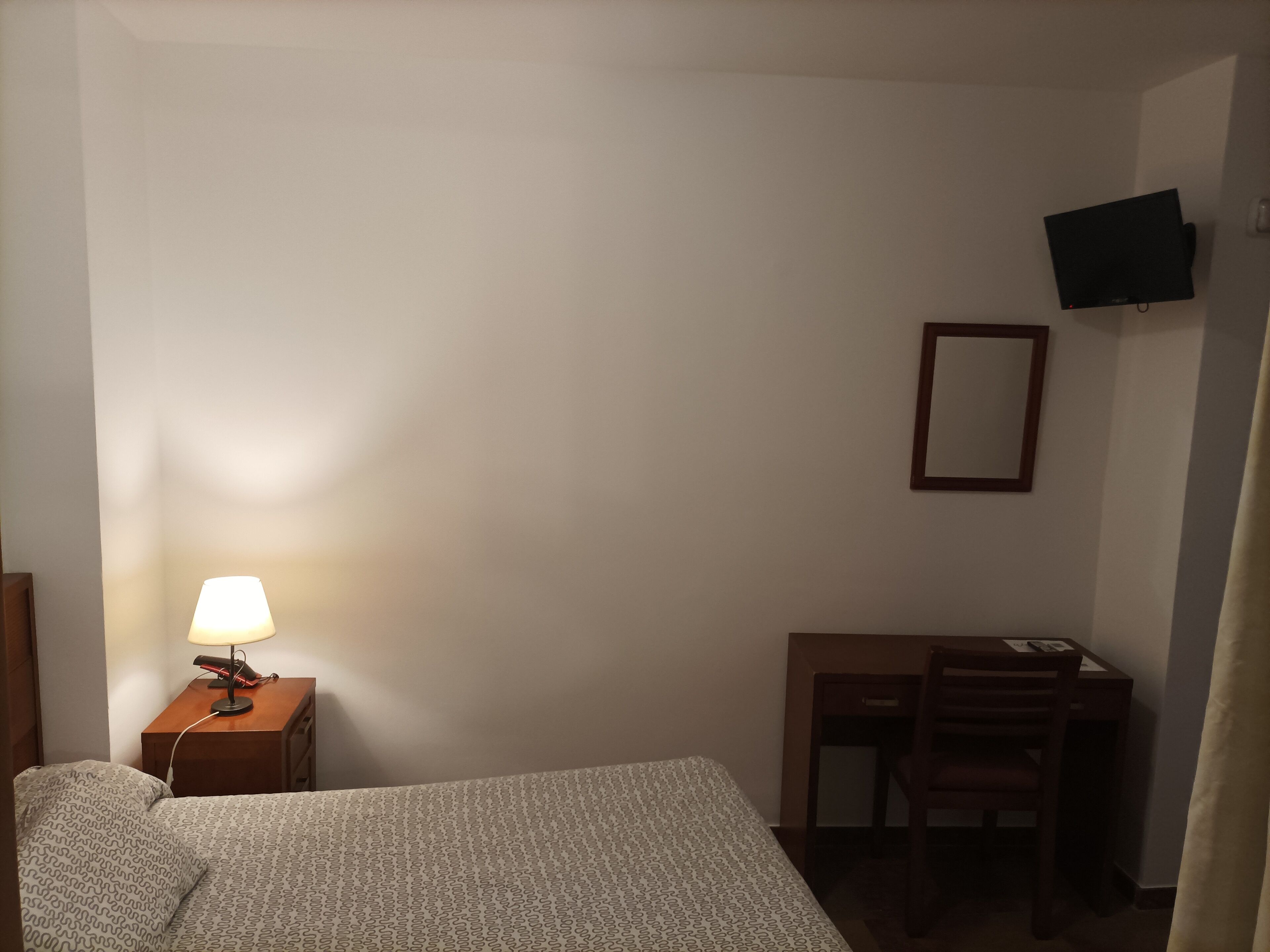 A bedroom with a bed, a desk, a chair, a lamp, and a TV mounted on the wall.