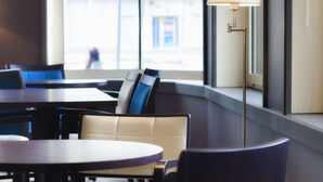 A modern indoor seating area with round tables and cushioned chairs, large windows with blinds, and a floor lamp. - Le Berry (Saint-Nazaire)