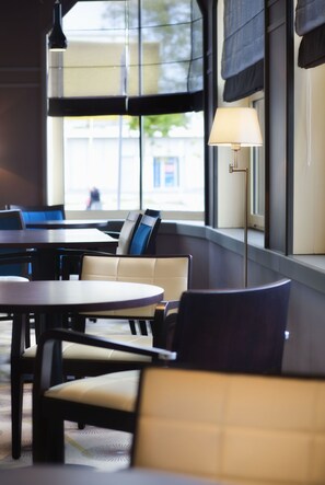 A modern indoor seating area with round tables and cushioned chairs, large windows with blinds, and a floor lamp. - Le Berry (Saint-Nazaire)