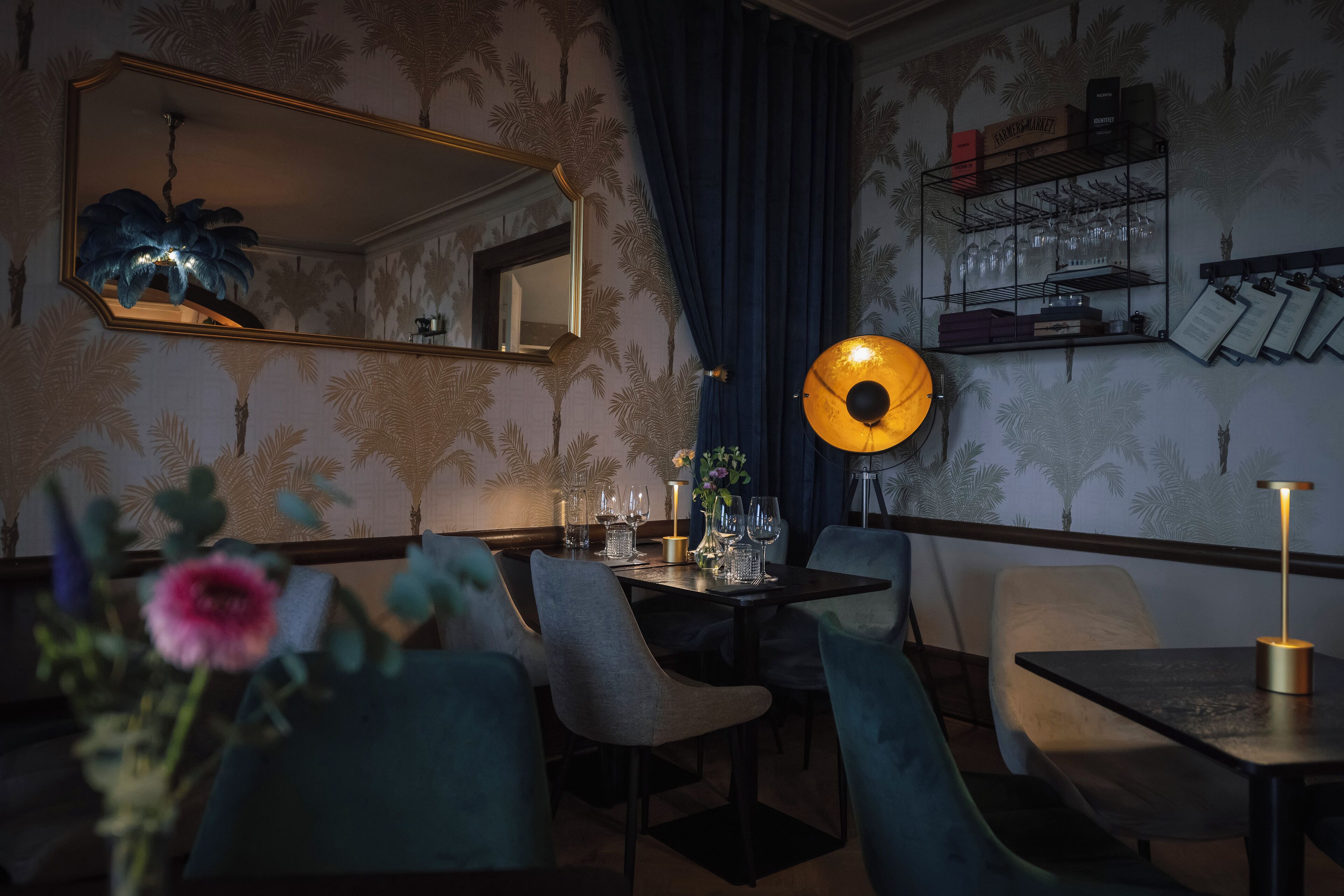 Comedor con una mesa dispuesta para dos, un espejo y un estante en la pared con vasos y documentos.