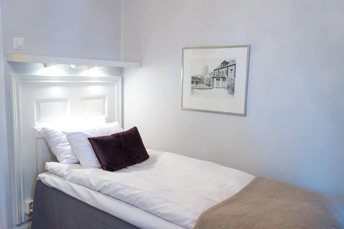 A neatly made bed with white linens and a brown pillow, a framed black and white photo on the wall, and a white headboard with built-in lighting.