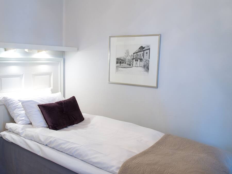 A neatly made bed with white linens and a brown pillow, a framed black and white photo on the wall, and a white headboard with built-in lighting.