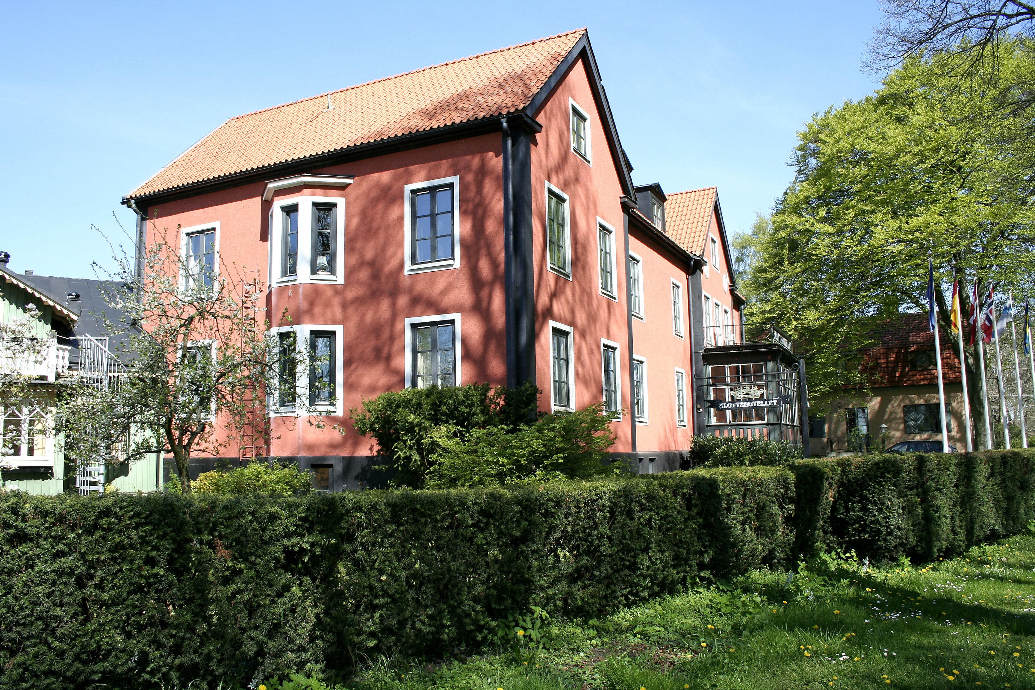 A red building with multiple windows and a tiled roof, surrounded by greenery and other buildings.