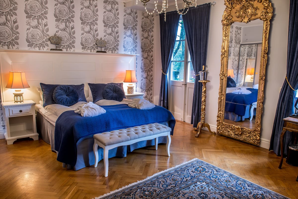 A bedroom with a large bed, two bedside tables with lamps, a bench, a large mirror, and a patterned rug.