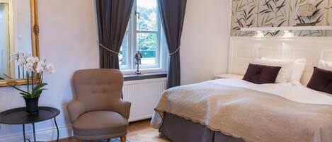 A bedroom with a bed, a chair, a small table, a mirror, and a window with curtains.