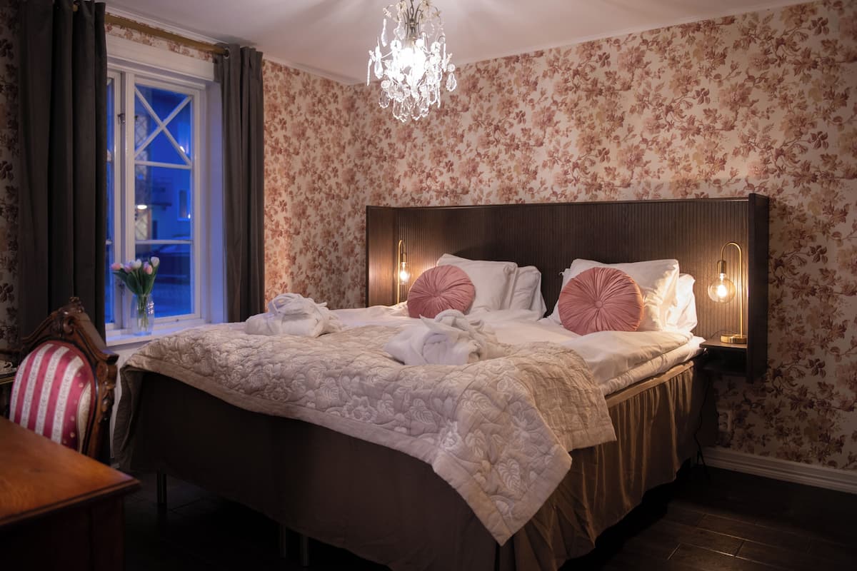 A hotel room with a large bed, floral wallpaper, a chandelier, and a window with curtains.
